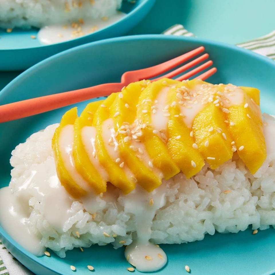 Thai mango sticky rice dessert with coconut milk and sesame seeds on a blue plate.
