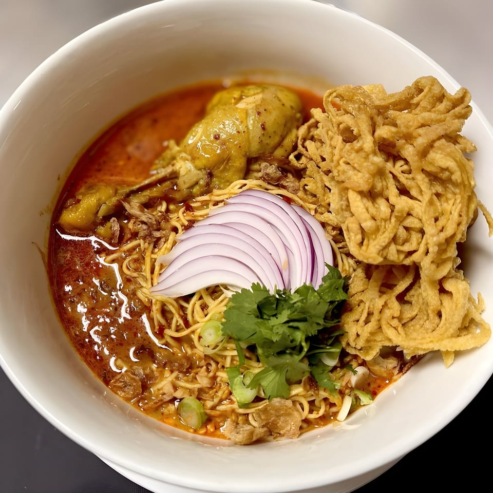 Close-up of a bowl of Thai Khao Soi with chicken, noodles, red onion, and crispy noodles.